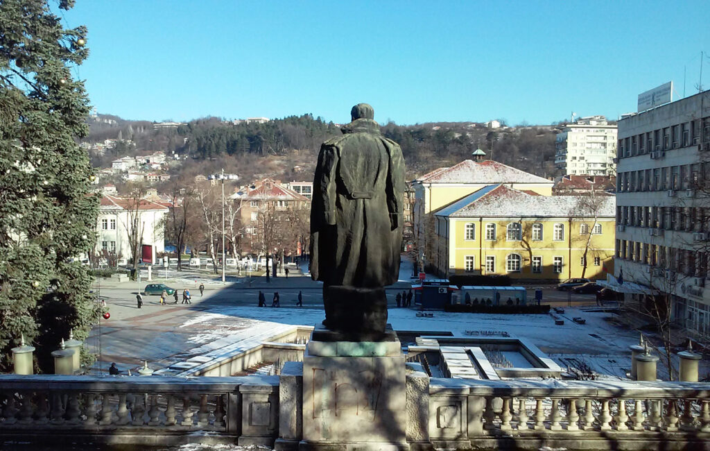 Pomnik Wasyla Lewskiego w Gabrowie, Bułgaria. Widok jest od tyłu, a w tle widać centralny plac miasta, otoczony charakterystycznymi budynkami, w tym żółtym budynkiem, który jest historycznym obiektem w Gabrowie.