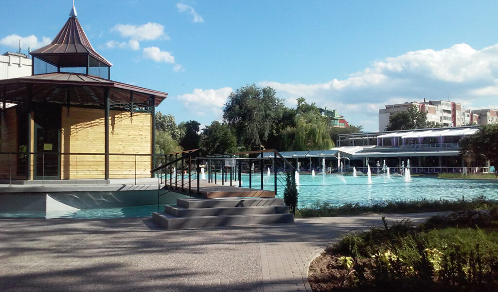 Nowoczesny kompleks AquaLand w Płowdiwie z altaną, basenem i fontannami, otoczony zielonym parkiem w słoneczny dzień.