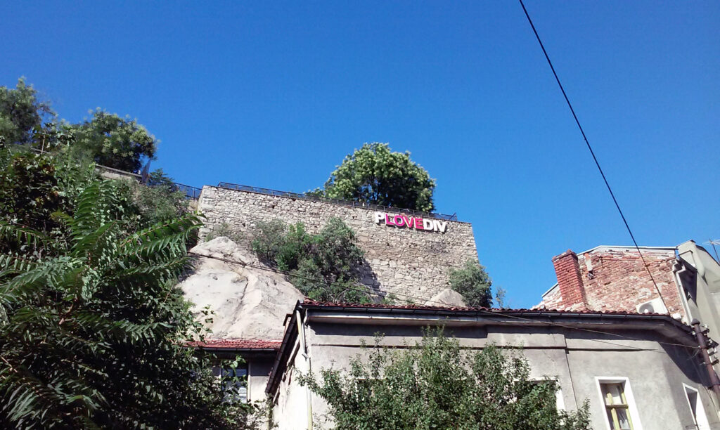 Wzgórze Nebet Tepe w Płowdiwie z napisem „PLOVEdiv” na kamiennych murach, otoczone zielenią, z widokiem na miasto i błękitne niebo.